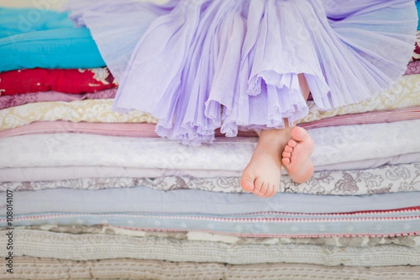 Fototapeta The fairy tale "the princess on a pea". Little girl in lilac tatu skirt sitting on the high bed. Bare foot if the girl. Legs of a little girl sitting on a pile of colorful mattresses.