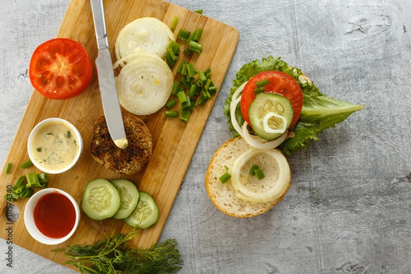 Fototapeta Cooking process of a sandwich burger, ingredients on wooden cutting board on wooden table against white background, fresh vegetables, herbs, fried meat, buns, sauces and knife