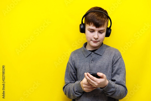 Fototapeta Boy in black wireless headphones looking at smartphone on yellow background.