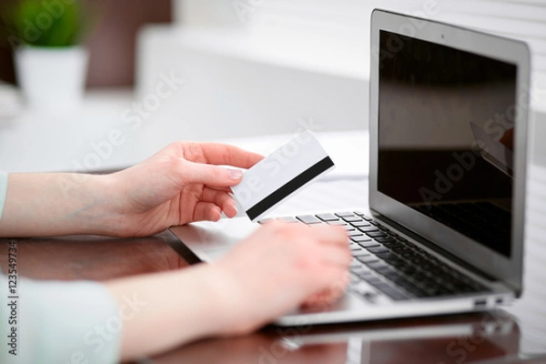 Fototapeta Business woman in a green blouse sitting at the desk in the office and working on laptop computer with a credit card in her hands .