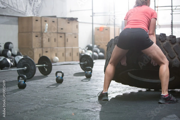 Fototapeta Rear view of woman flipping tire in crossfit gym