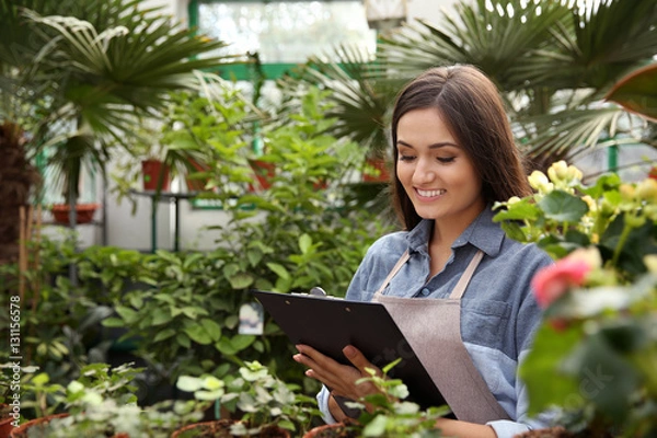 Fototapeta Pretty young florist looking after decorative plants in greenhouse