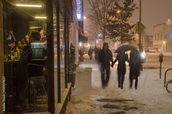 Fototapeta Customers in a bar are seen through a window while pedestrians walk through the snow outdoors.