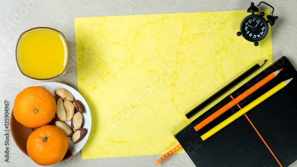 Fototapeta Black notebook, sheet of handmade yellow paper, color pencils, clock, glass of orange juice and healthy snack on light wooden background. Colorful work space. Desk dining.Top view. Ratio 16:9.