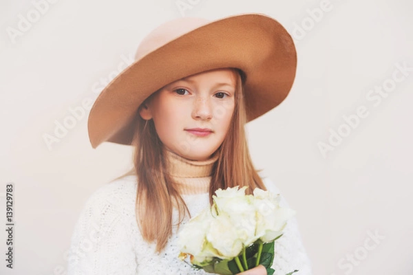 Fototapeta Pretty little girl wearing a hat, holding bouquet of white roses, standing against beige background