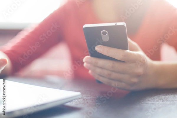 Fototapeta Cropped shot of female hand holding her smartphone in hand reading message via email or making call sitting at wooden table  and working with her laptop. Film effect. Technology concept.