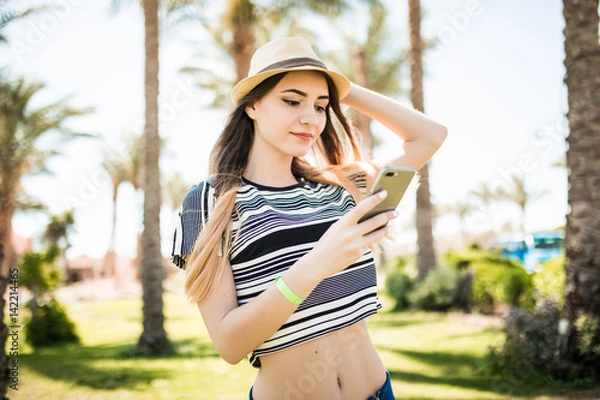 Obraz Happy beauty young woman in hat walking and using smartphone on summer resort