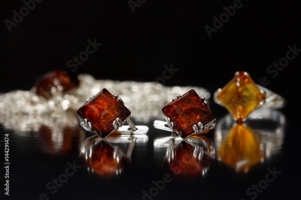 Fototapeta Closeup silver earrings rhombus with authentic natural baltic amber on black  surface on background of ring and chain with pendant.