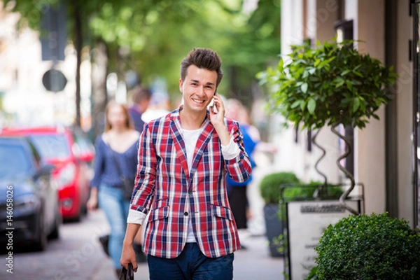 Fototapeta Portrait of handsome smiling stylish man with bag and smart phone in plaid blazer in the summer city. Talking on the phone. Street style concept. Walking down the sunny city street. 