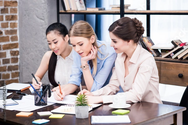 Fototapeta Multiethnic group of young businesswomen working together at table with papers