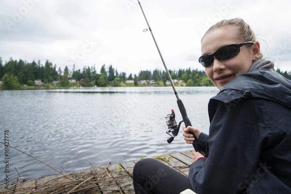 Fototapeta   Teenage girl fishing in a lake while sitting on a dock