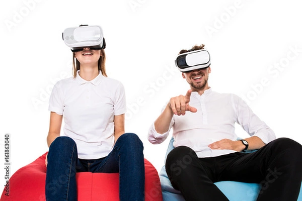 Fototapeta Excited young couple experiencing virtual reality seated on beanbags isolated on white background