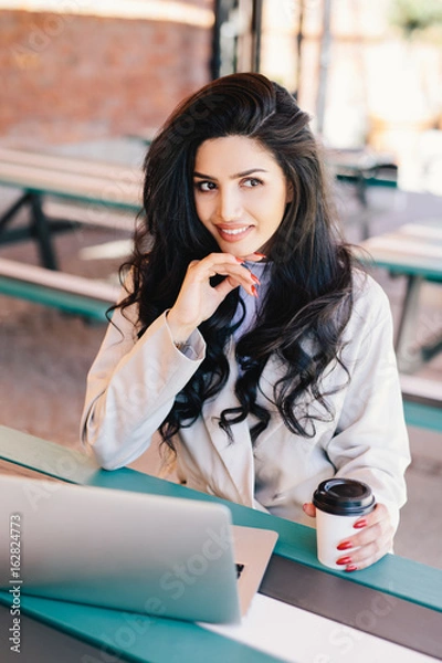 Fototapeta Caucasian young female freelancer with long wavy hair having dark eyes and healthy skin looking mysteriously aside working remotely on modern laptop pc, using free wi-fi drinking tasty coffee.