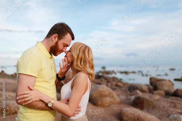 Fototapeta Young beautiful smiling couple hugging on the shore amid rocks and sea, enjoy the sunset and freedom. Male to prepare for a kiss. Journey to the sea.