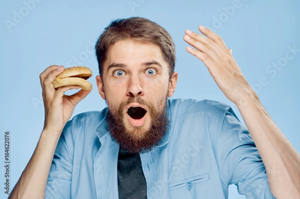 Fototapeta Young guy with a beard on a blue background holds a hamburger