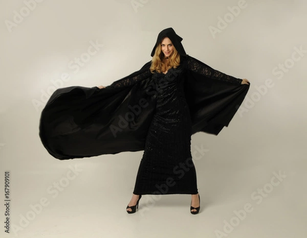 Fototapeta full length portrait of a blonde lady wearing black lace now and hooded cloak, standing pose isolated against creamy background.