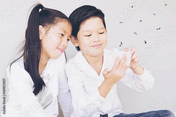 Fototapeta Lovely Asian couple school kids are playing song on mobile phone, 7 and 10 years old, over gray background