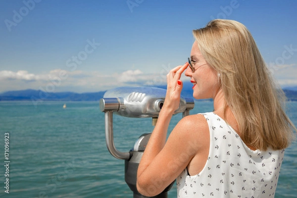 Fototapeta young sexy woman enjoy the beautiful view with a coin operated binoculars. The sun is shining, the water and the sky is blue. she wear sunglasses an a white dress