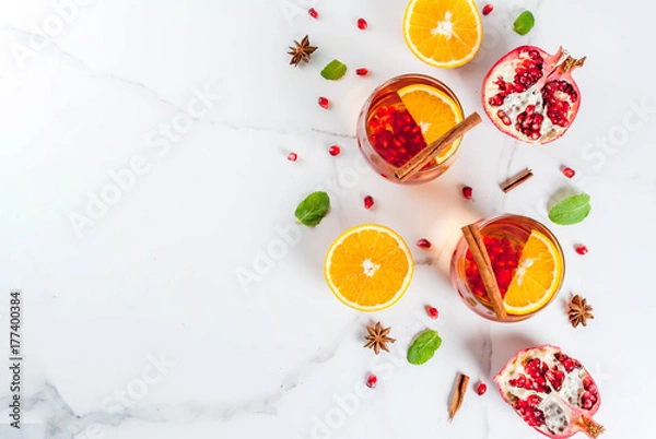 Fototapeta Fall and winter drinks. Warm refreshing cocktail with  pomegranate, oranges, cinnamon, spices and mint. On a white marble table. Copy space  top view