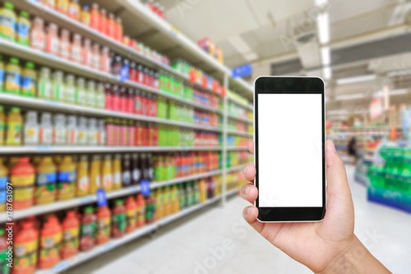 Fototapeta Hand holding smartphone with white blank screen and blurred supermarket drink background