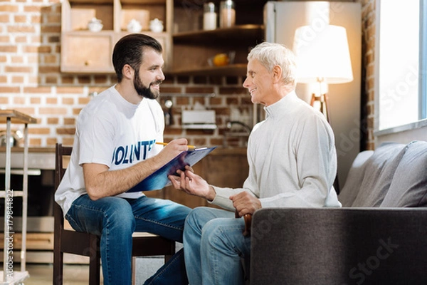 Fototapeta Writing everything. Enthusiastic young social worker looking happy while sitting with a great senior man and writing important information about him