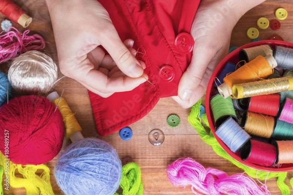 Fototapeta Woman`s hand holding a needle and colorful sewing and knitting supplies on the wooden table, bowl with bobbins, knitting yarn and buttons.