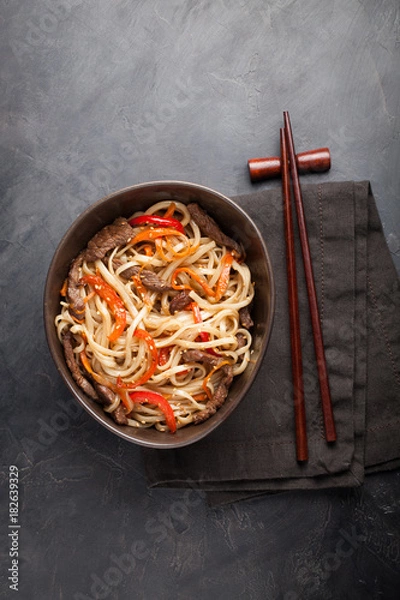 Fototapeta Close-up of noodles in a glass bowl with vegetables and beef in teriyaki sauce on a dark concrete background. Top view of Asian food, and wooden sticks. The concept of fast food