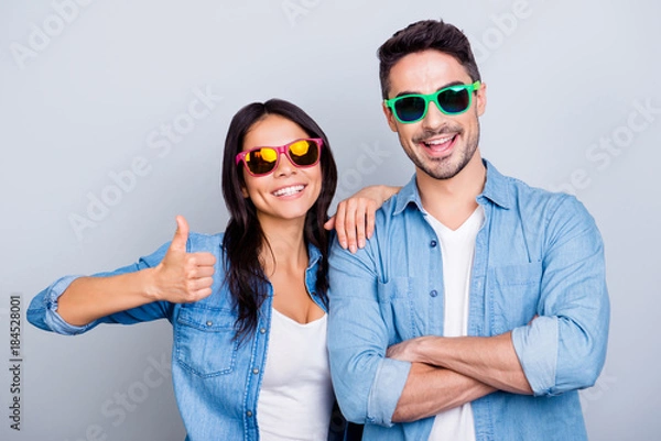 Fototapeta Attractive cool beautiful partners in summer glasses smiling to the camera, girl making thumb up sign, man with crossed hands over grey background