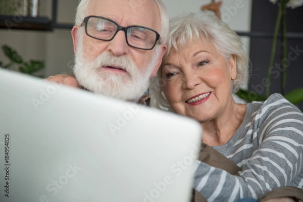 Fototapeta Close up portrait of smiling pensioners talking with family through computer. Focus on woman putting hand on male shoulder