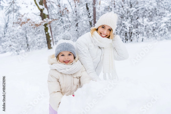 Fototapeta beautiful happy mother and daughter smiling at camera while making snowman in winter park