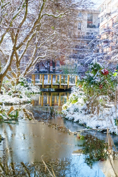 Fototapeta Small river with mini pont in the park, outdoor in Paris, France, during snowfall in winter. Taken in the morning with sunrise light. Smart concept of modern park for family recreation.