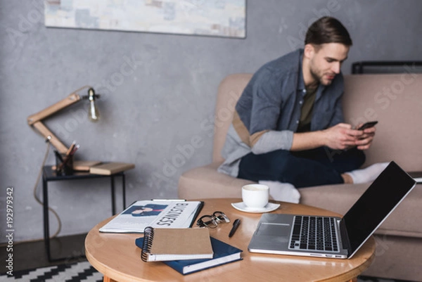 Obraz young handsome man using smartphone on couch with laptop and business supplies standing on table on foreground