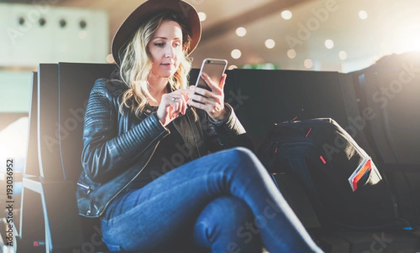 Fototapeta Young woman tourist in hat, with backpack sits at airport and uses smartphone. Hipster girl is waiting for plane landing, checks email, chatting, blogging, browsing internet.