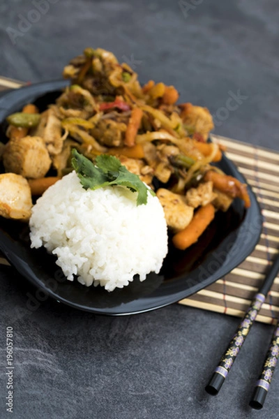 Fototapeta Fried vegetables with turkey meat and rice on the black plate. Asian delicates dishes with chili sauce.