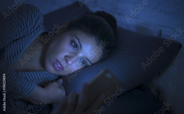 Fototapeta portrait of young beautiful latin woman using mobile phone late night sleepless lying in bed in the dark in smartphone and internet dating app addiction