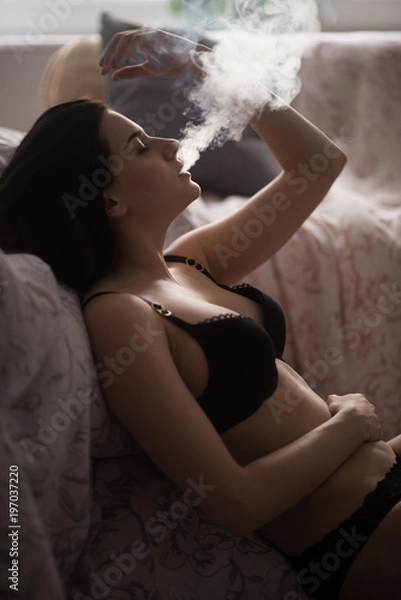 Fototapeta Sexy young woman in a black lingerie vaping in bedroom. Beautiful brunette woman smoking