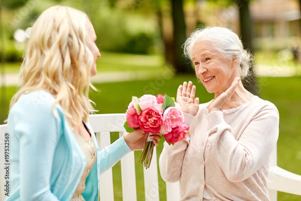 Fototapeta family, holidays and people concept - happy smiling young daughter giving flowers to her senior mother sitting on park bench