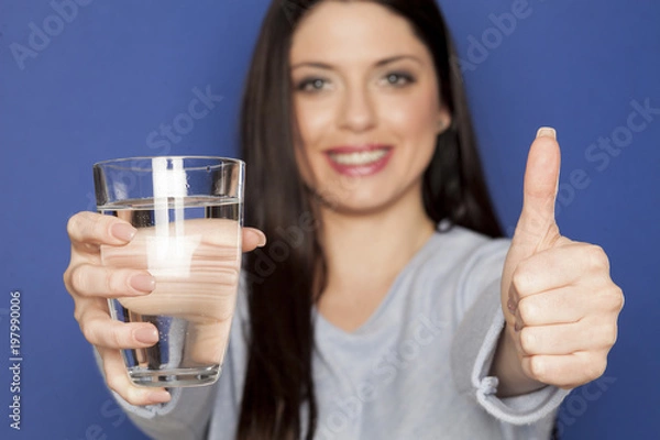 Fototapeta A young lady holding a glass with wather on a blue background and showing thumbs up