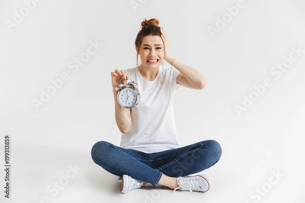 Fototapeta Portrait of a confused young girl holding alarm clock