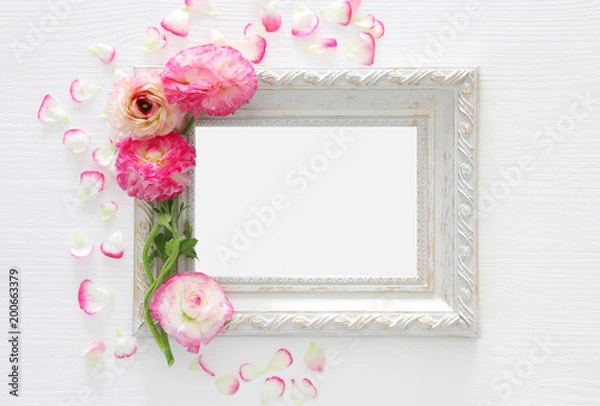 Fototapeta Image of delicate pastel pink beautiful flowers arrangement and empty vintage photo frame over white wooden background. Flat lay, top view. For photography mockup montage.