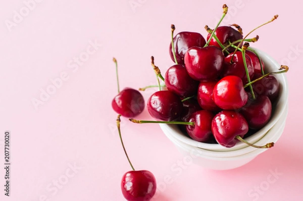 Fototapeta Heap of fresh ripe red cherries in a white bowl on a light pink background. Top view and copy space. Organic food concept.