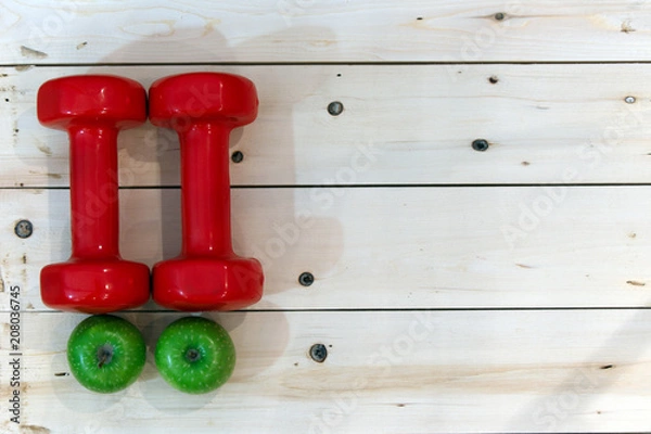 Fototapeta two red dumbbells along with two green apples on white wooden background. Copy space. concept