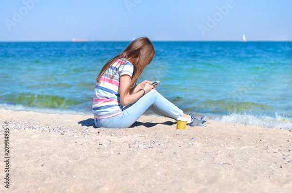 Fototapeta Young beautiful woman enjoys relaxing at the sea. She sits on the sand on a blue sea background with a smartphone in her hands