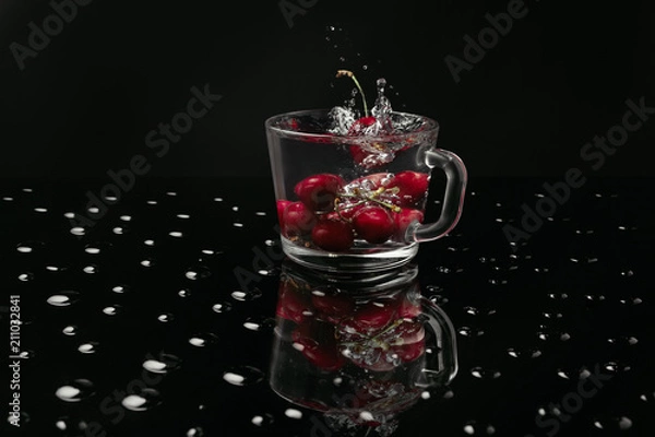 Fototapeta Clear glass mug with cherries in front of a black background on a black table made of glass covered with water drops. Reflection of a mug with cherries. Falling berries create splashes of water.