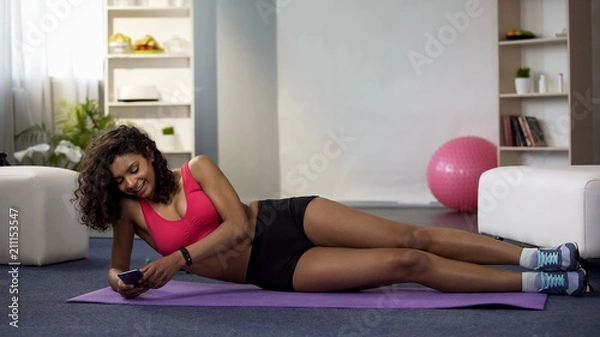 Fototapeta Mixed race female lying on floor in sportswear, using phone, procrastination