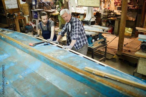 Fototapeta Young and senior shipbuilding workers standing by large vessel construction and repairing it