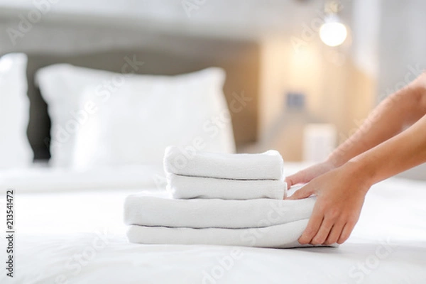 Fototapeta Close-up of hands putting stack of fresh white bath towels on the bed sheet. Room service maid cleaning hotel room.