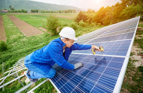 Fototapeta Construction worker connects photo voltaic panel to solar system using screwdriver. Professional installing and construction of solar system, alternative energy and financial investment concept.