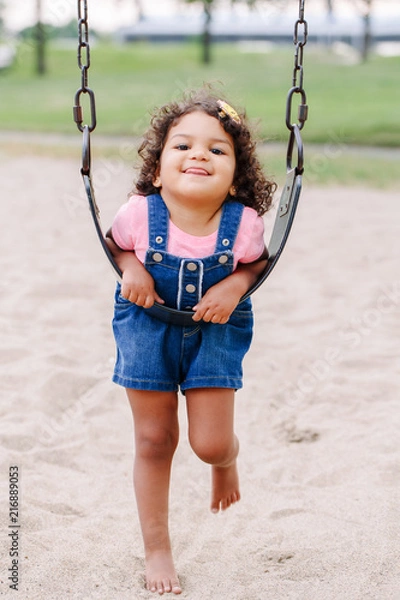 Fototapeta Portrait of happy smiling little latin hispanic toddler girl swinging on swings at playground outside on summer day. Happy childhood lifestyle concept. Toned with film pastel faded filters colors.