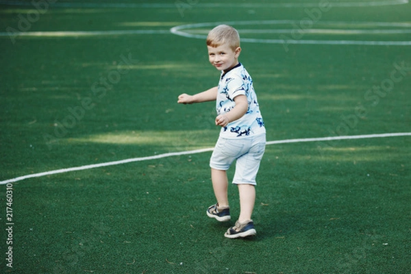 Fototapeta Happy cute blond boy in blue t-shirt and shorts on a football field without a ball.